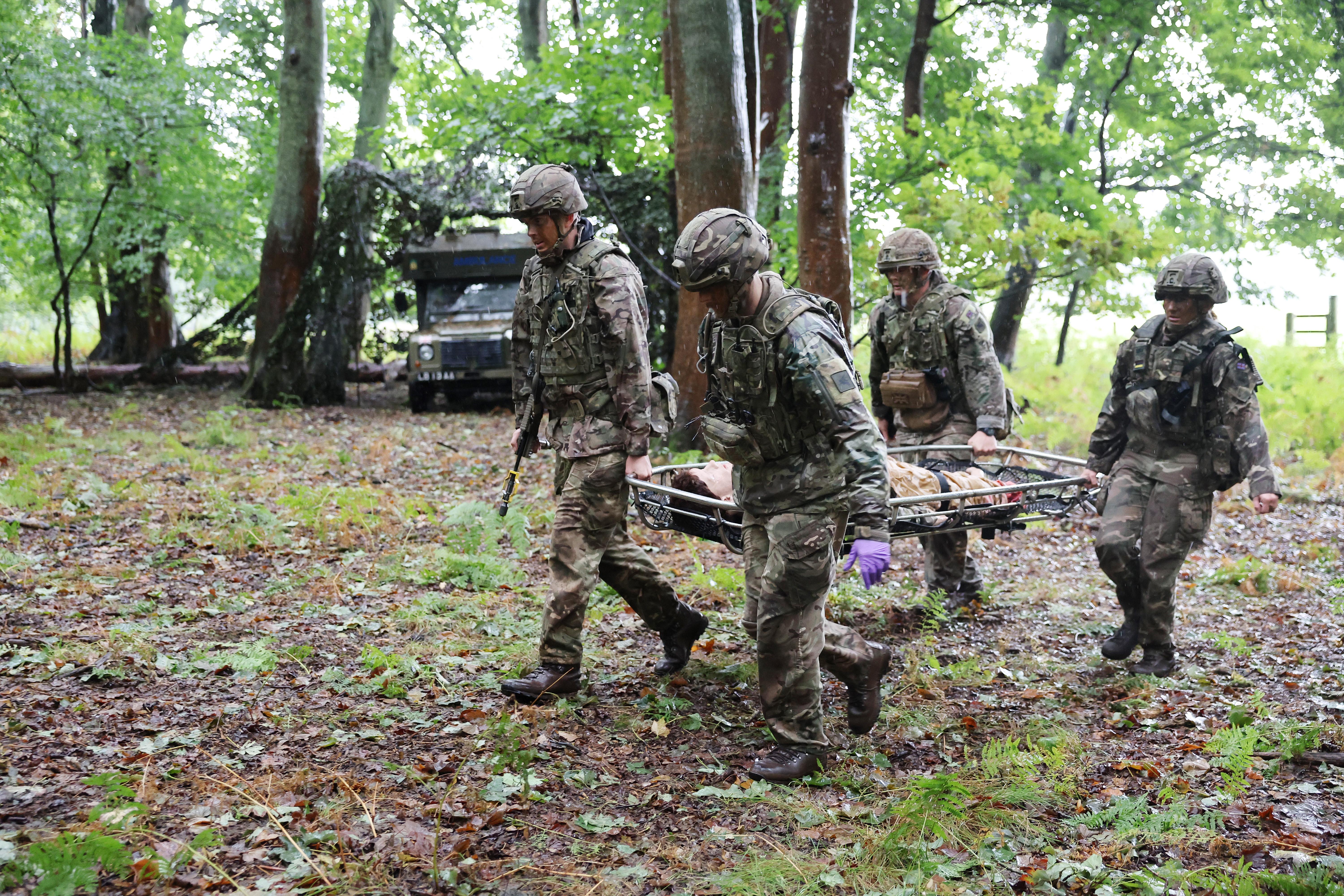 Soldiers in camouflage uniforms and helmets are carrying a stretcher through a wooded area.