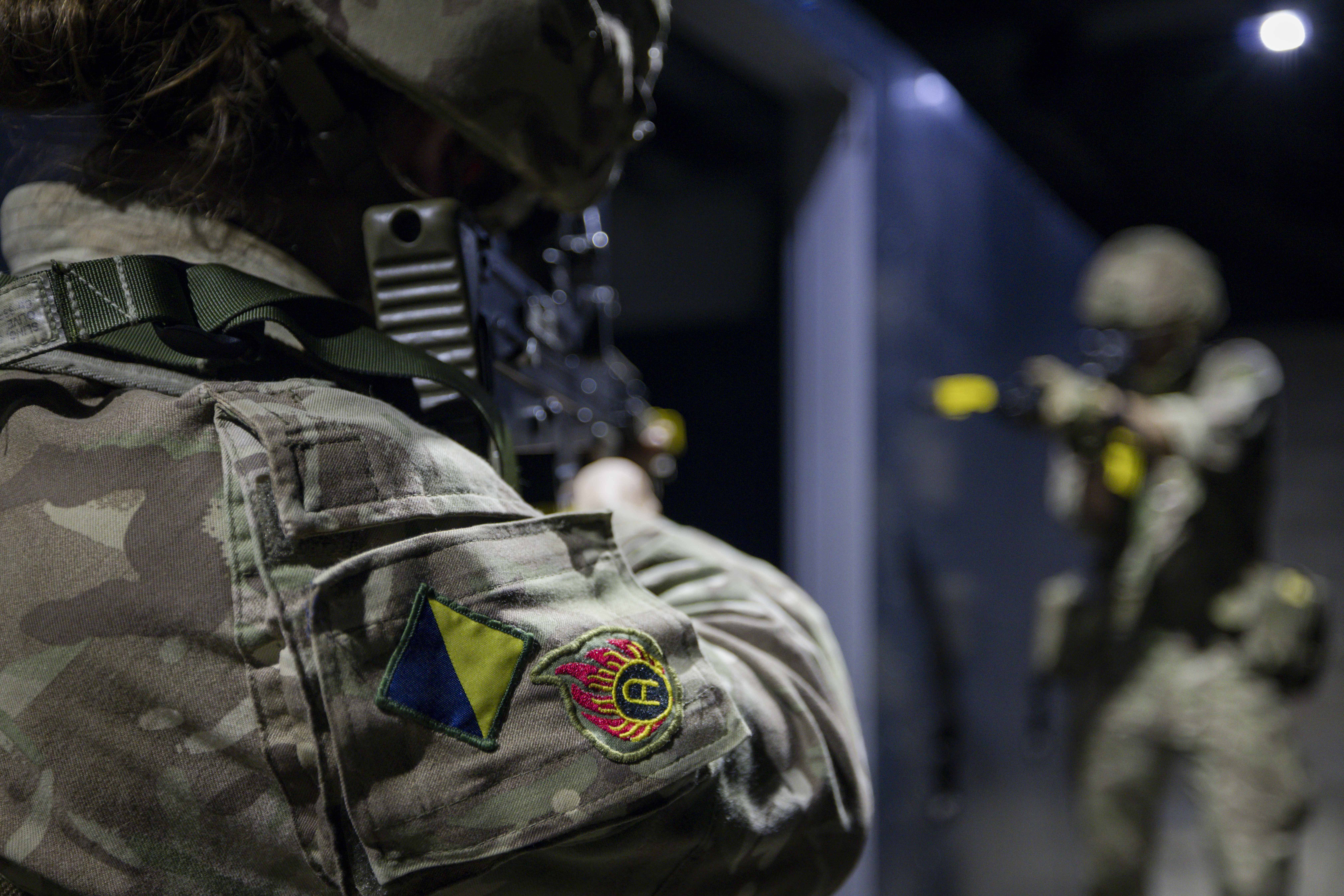 Military personnel in camouflage uniforms participate in a training exercise. One person holds a firearm and faces another individual, who is also armed. The environment appears to be indoors with dim lighting.