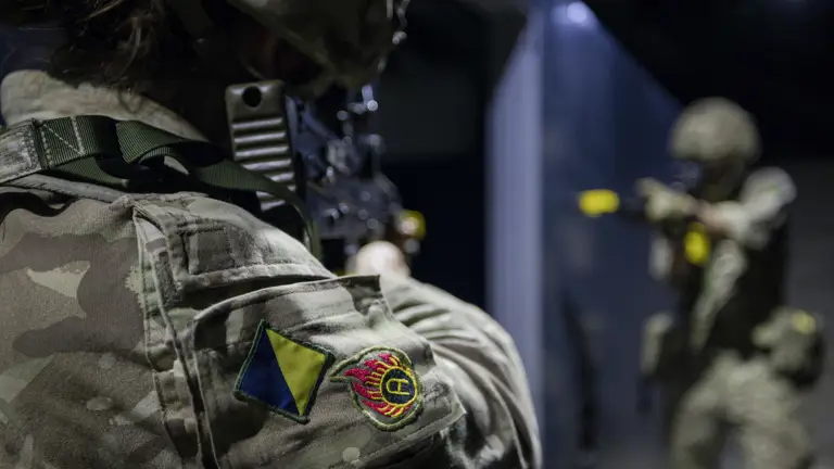 Military personnel in camouflage uniforms participate in a training exercise. One person holds a firearm and faces another individual, who is also armed. The environment appears to be indoors with dim lighting.