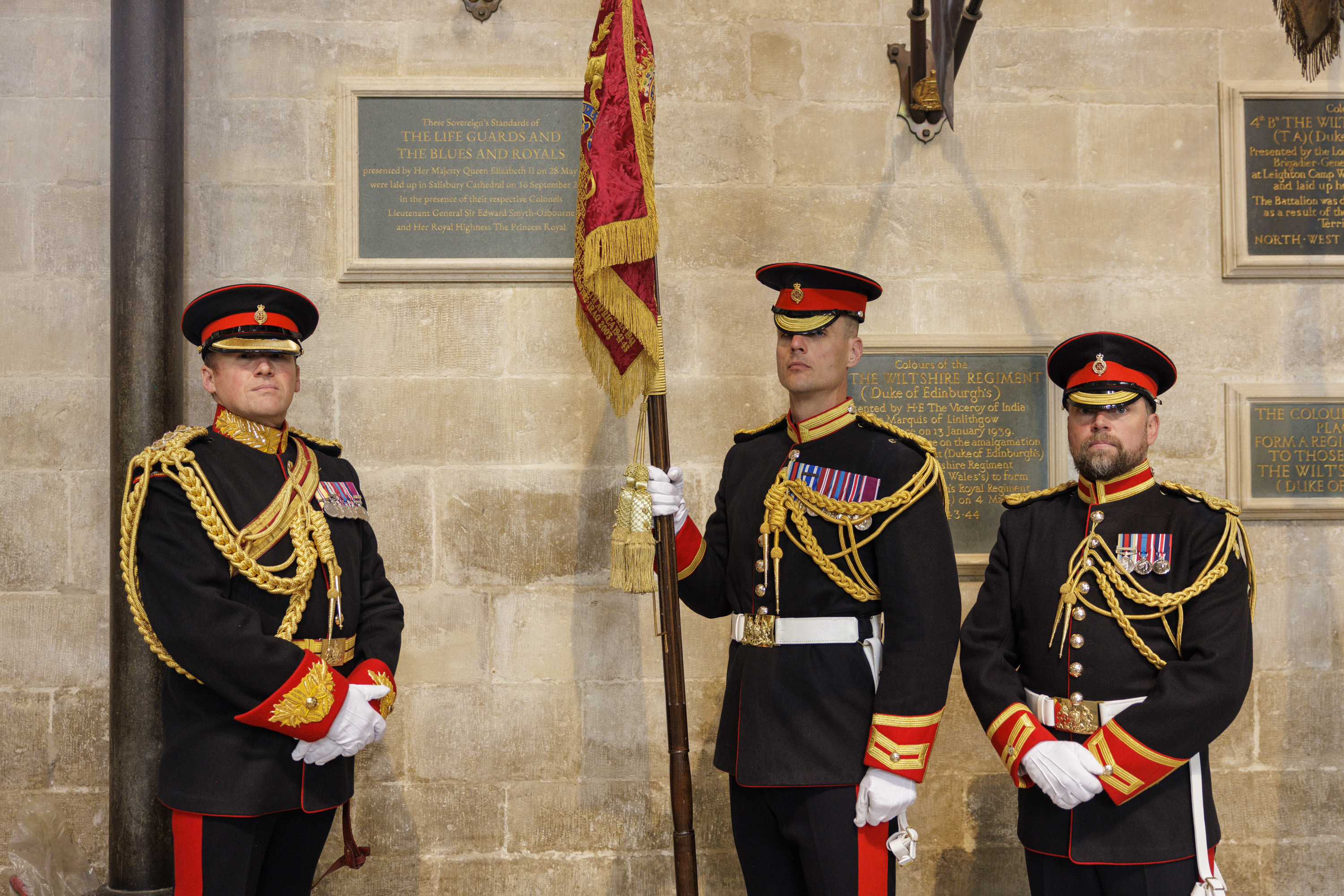 Three men in black military ceremonial uniforms stand for a portrait with their red and gold ceremonial flag.