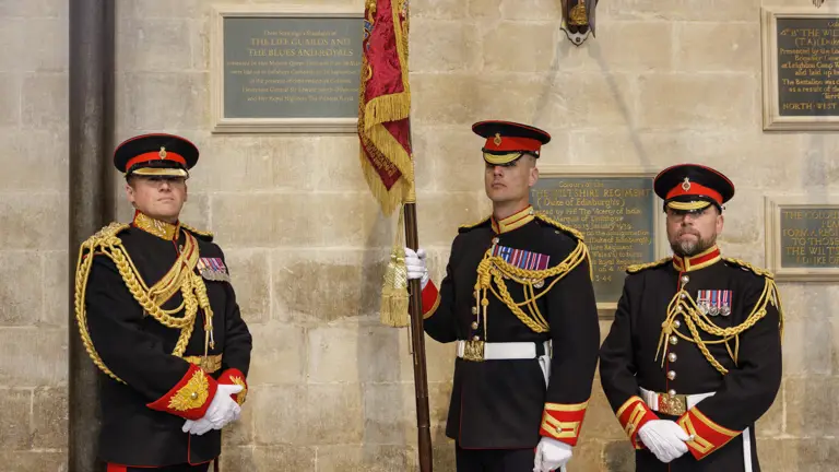 Three men in black military ceremonial uniforms stand for a portrait with their red and gold ceremonial flag.
