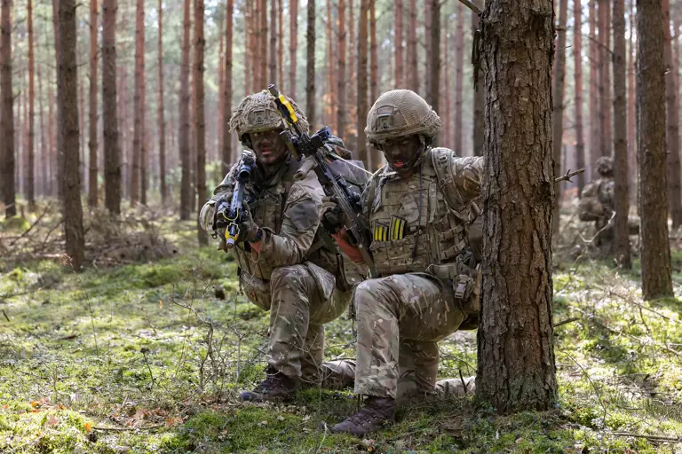 Two soldiers in camouflage uniforms kneel behind a tree. They are holding rifles.