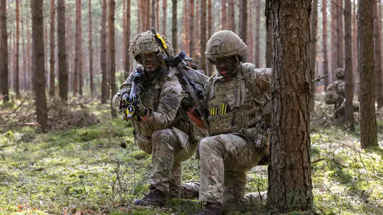 Two soldiers in camouflage uniforms kneel behind a tree. They are holding rifles.