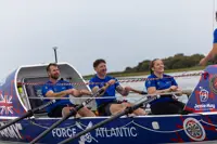 Three rowers in blue shirts rowing a Force Atlantic boat on calm water with a cloudy sky.