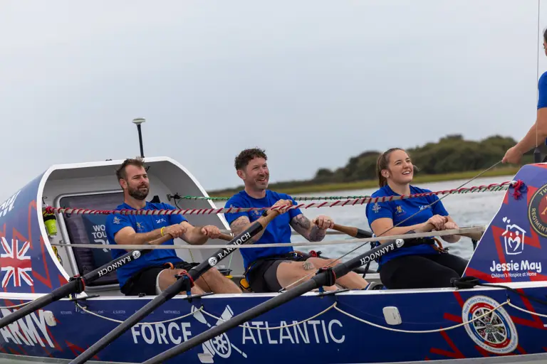 Three rowers in blue shirts rowing a Force Atlantic boat on calm water with a cloudy sky.