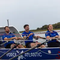Three rowers in blue shirts rowing a Force Atlantic boat on calm water with a cloudy sky.