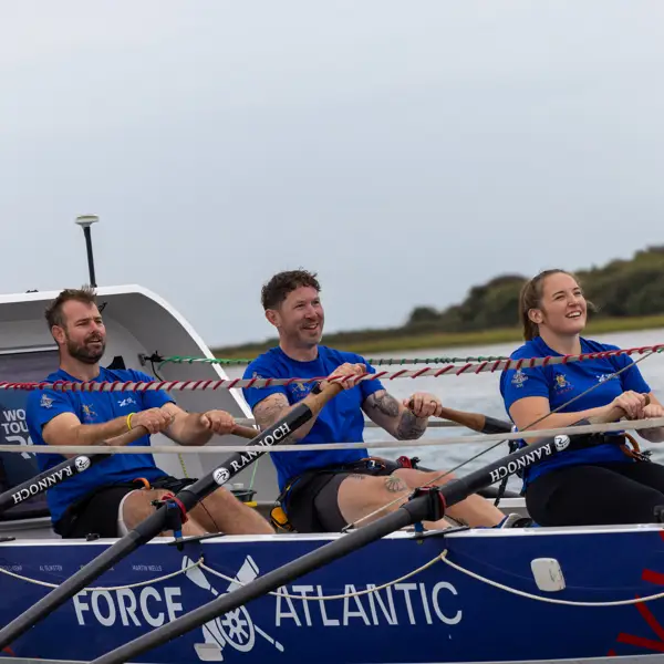 Three rowers in blue shirts rowing a Force Atlantic boat on calm water with a cloudy sky.