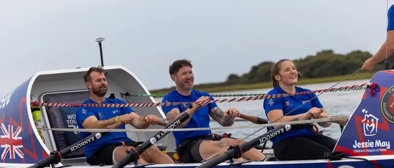Three rowers in blue shirts rowing a Force Atlantic boat on calm water with a cloudy sky.