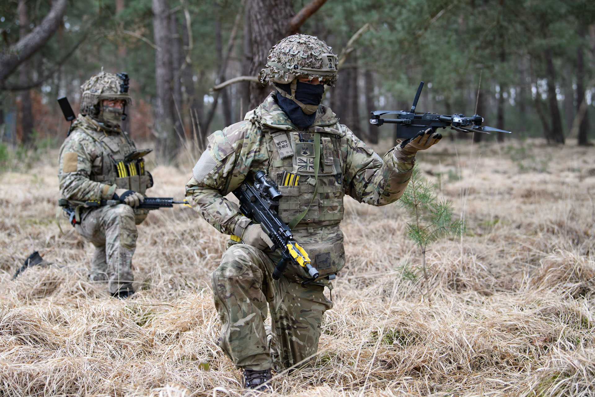 Two soldiers in camouflage gear are in a forest. One is kneeling and holding a small drone, while the other stands behind.