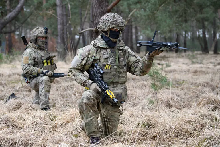 Two soldiers in camouflage gear are in a forest. One is kneeling and holding a small drone, while the other stands behind.