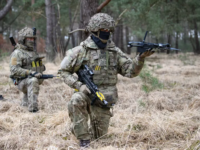 Two soldiers in camouflage gear are in a forest. One is kneeling and holding a small drone, while the other stands behind.