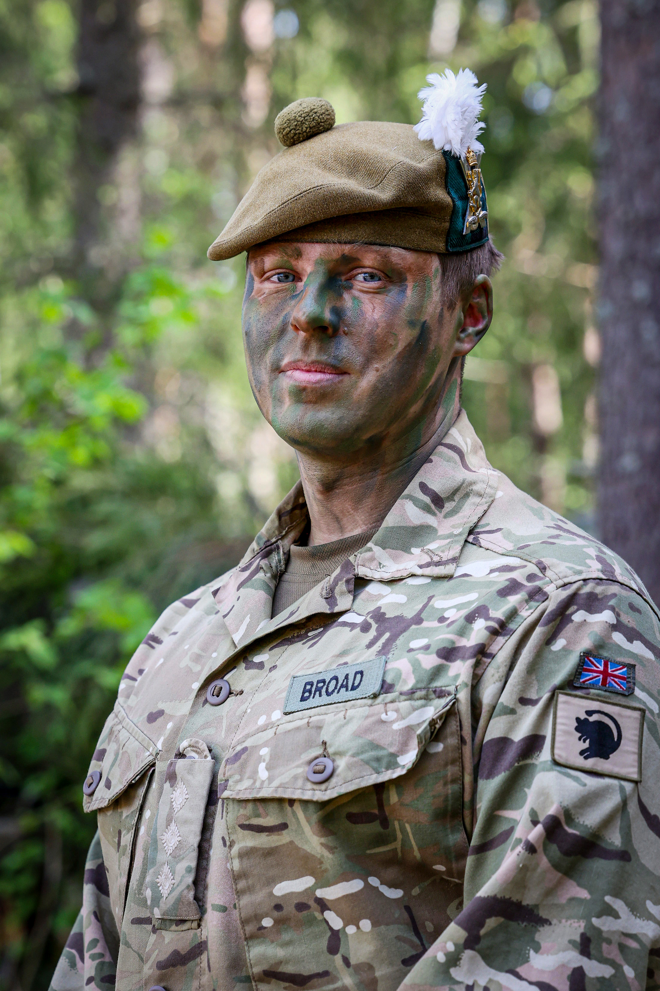  Military personnel in camouflage face paint and uniform stand in a forested area, with patches on the uniform.