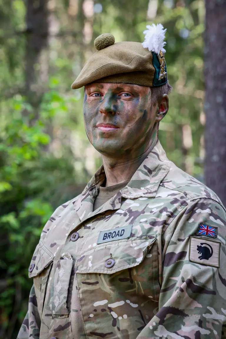 Military personnel in camouflage face paint and uniform stand in a forested area, with patches on the uniform.