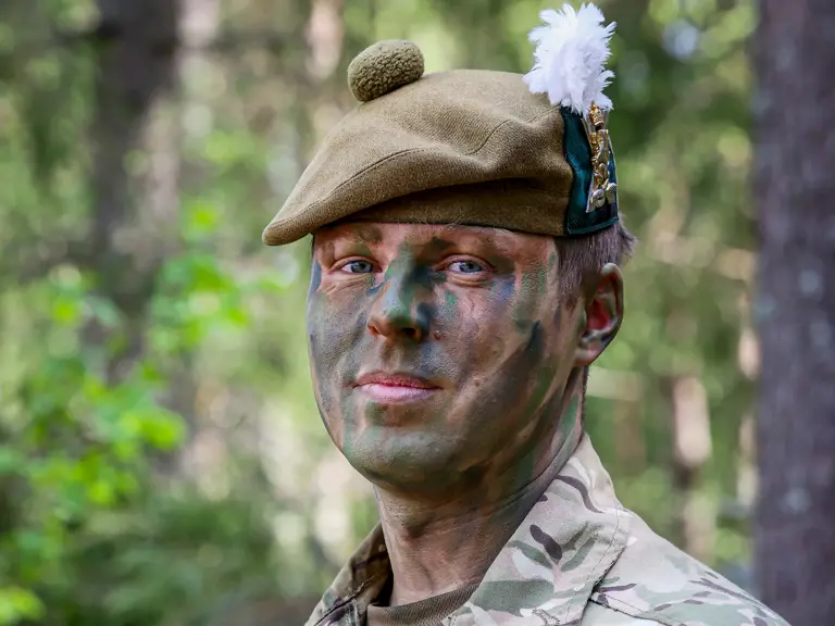 Military personnel in camouflage face paint and uniform stand in a forested area, with patches on the uniform.