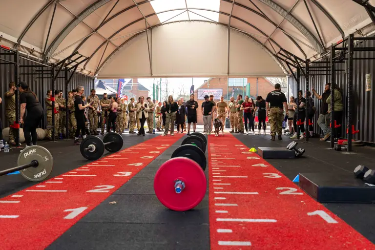 A CrossFit gym with equipment set on red numbered mats. People in military uniforms and athletic attire stand, watching in anticipation, creating a spirited atmosphere.