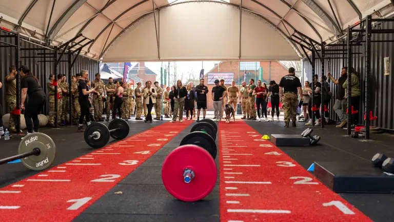 A CrossFit gym with equipment set on red numbered mats. People in military uniforms and athletic attire stand, watching in anticipation, creating a spirited atmosphere.