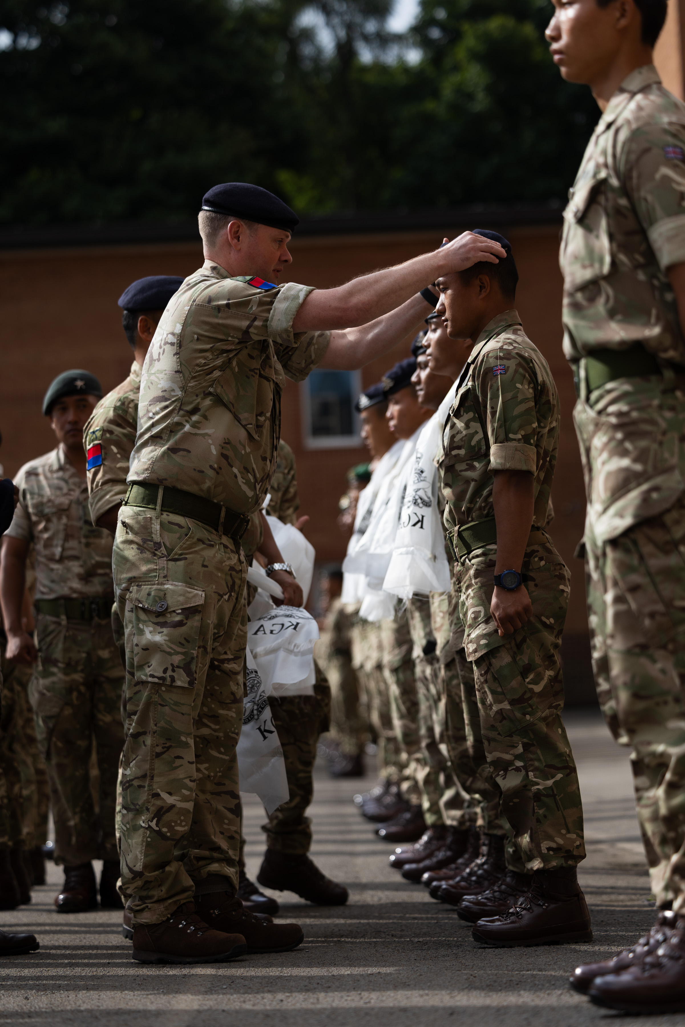 A British Army Officer wearing camouflage uniform, is placing a beret on the head of a Gurkha soldier, who is part of a line of soldiers receiving new cap badges.