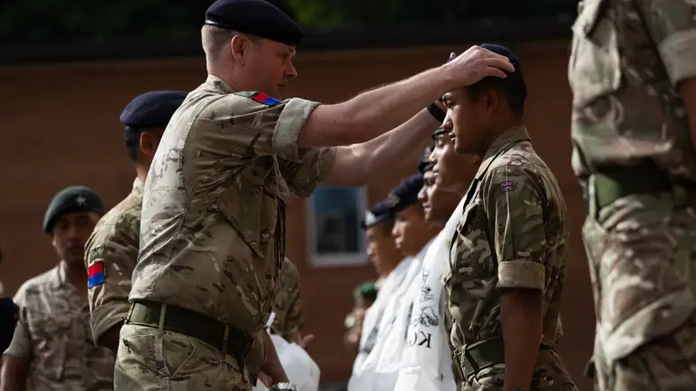 A British Army Officer wearing camouflage uniform, is placing a beret on the head of a Gurkha soldier, who is part of a line of soldiers receiving new cap badges.