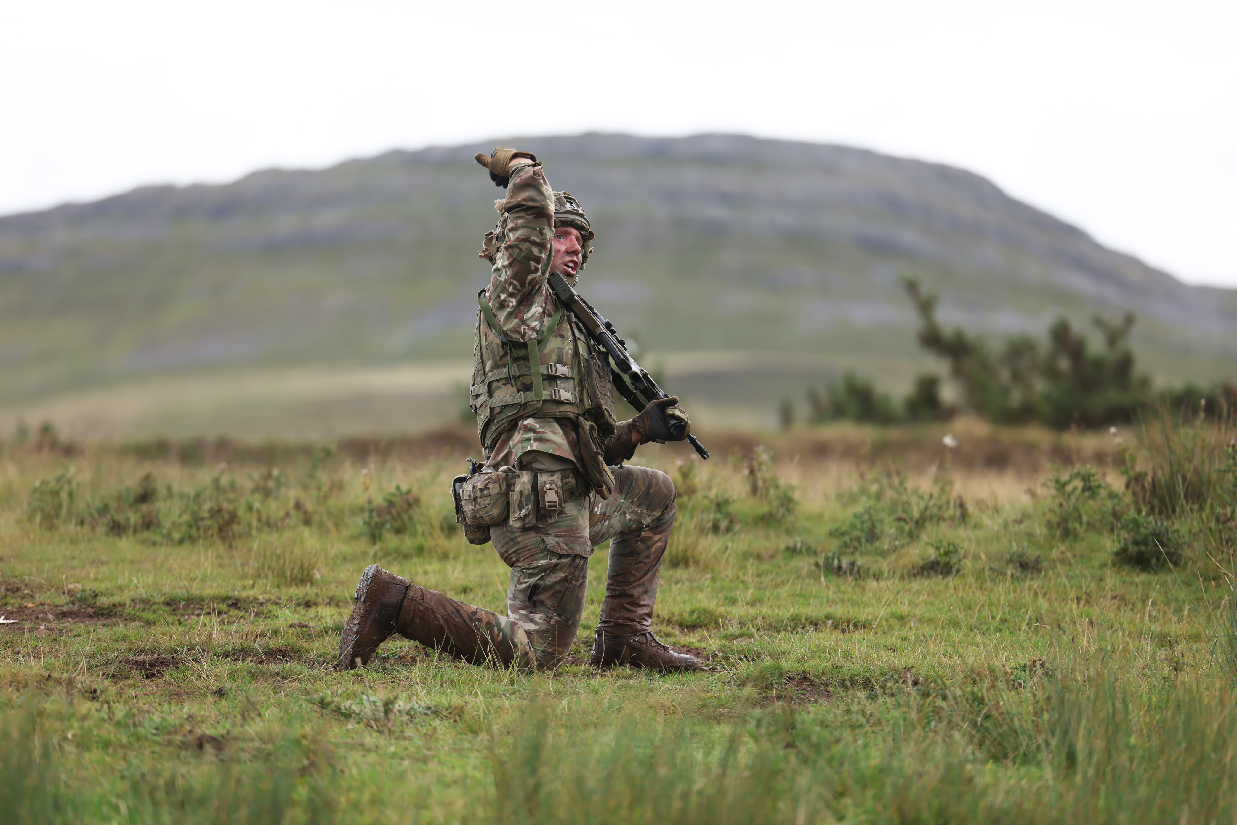 Soldier in full combat gear kneeling on grassy terrain with a rifle raised, set against a blurred mountainous background.
