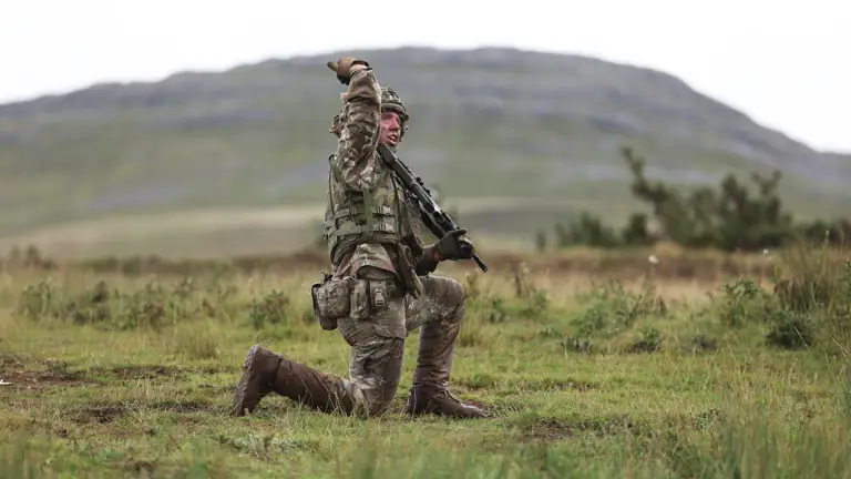 Soldier in full combat gear kneeling on grassy terrain with a rifle raised, set against a blurred mountainous background.