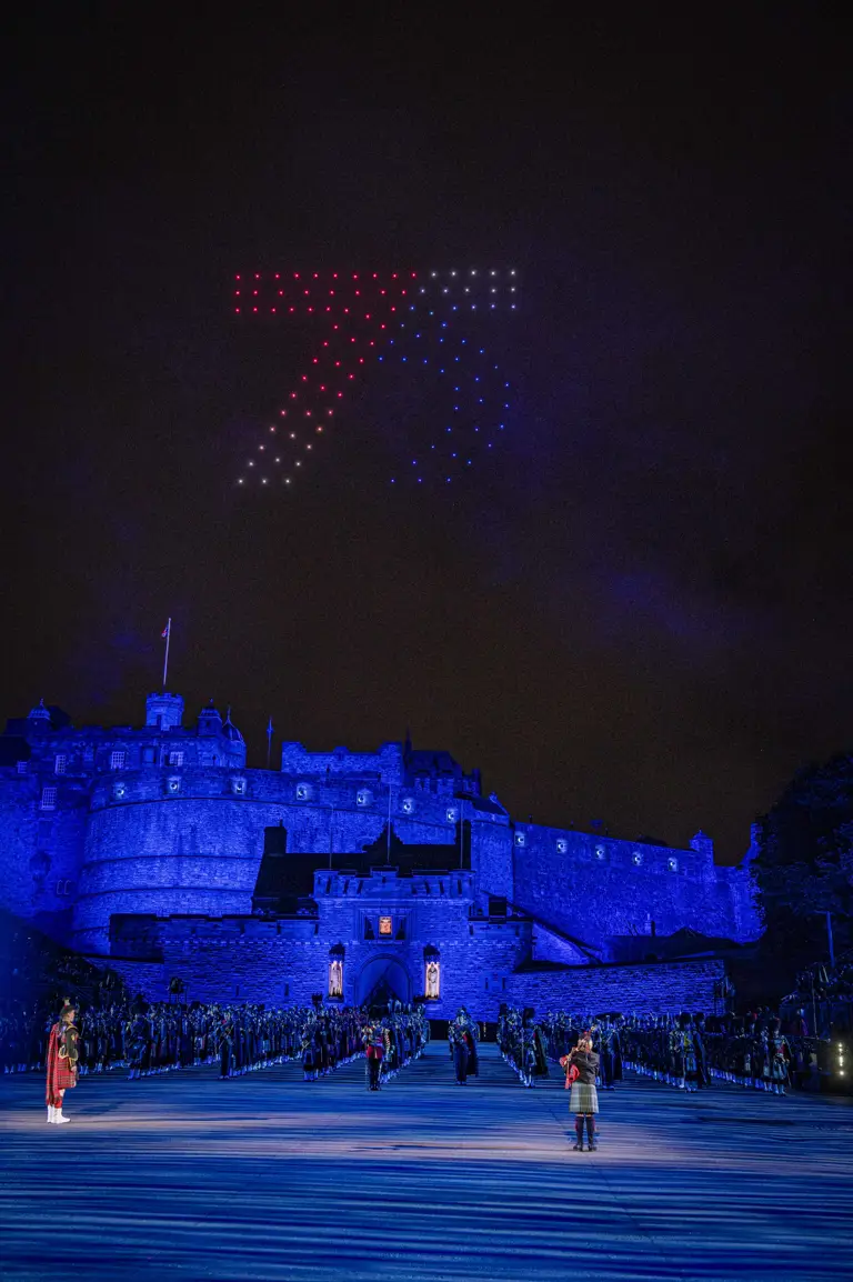 Pipers, drummers and musicians stood in line formation in front of a blue lit Edinburgh Castle Esplanade at night.