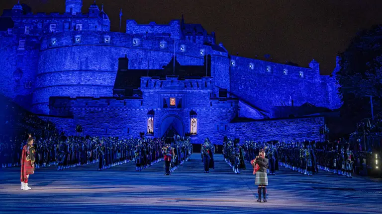 Pipers, drummers and musicians stood in line formation in front of a blue lit Edinburgh Castle Esplanade at night.