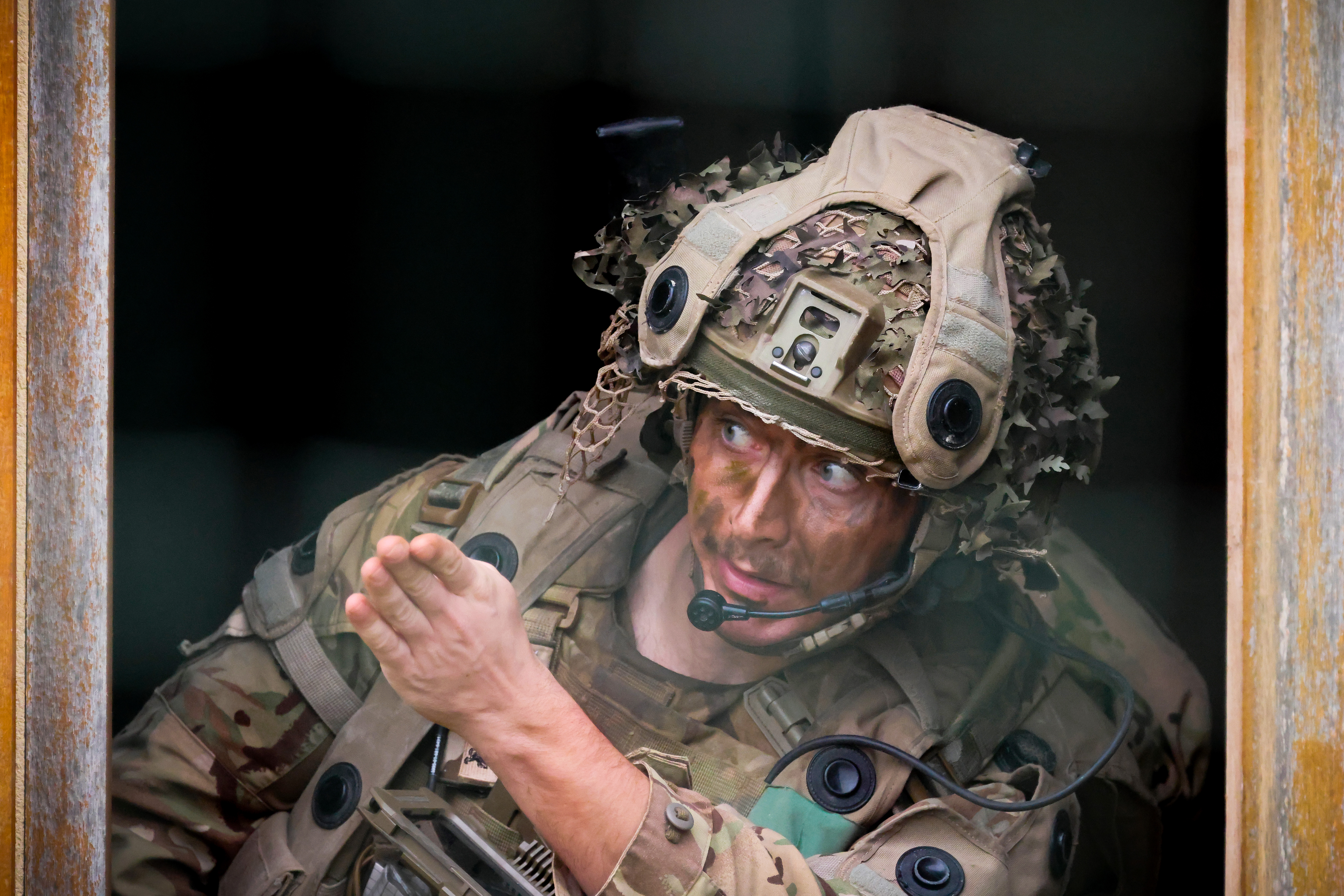 A soldier in camouflage uniform and helmet, equipped with a communications headset, gestures while looking ahead. 