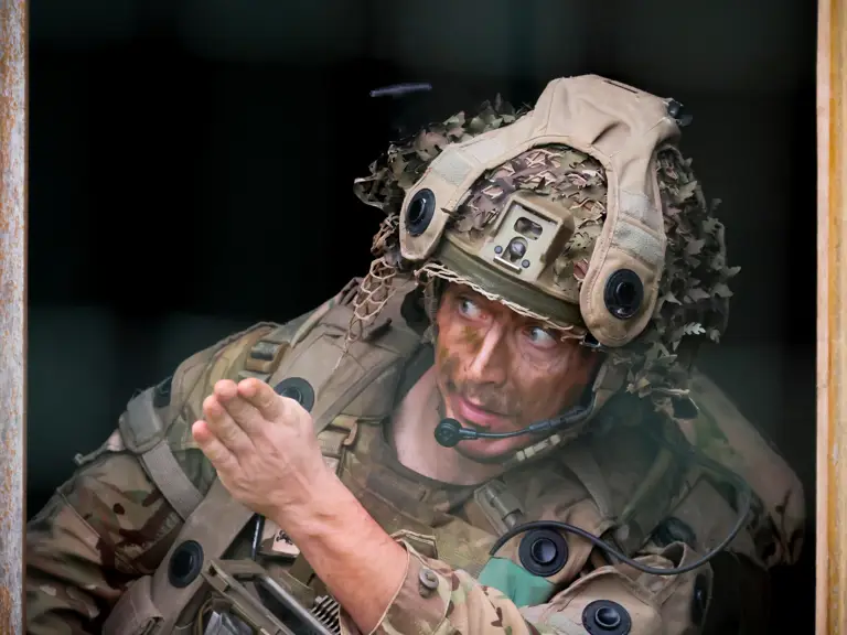 A soldier in camouflage uniform and helmet, equipped with a communications headset, gestures while looking ahead.