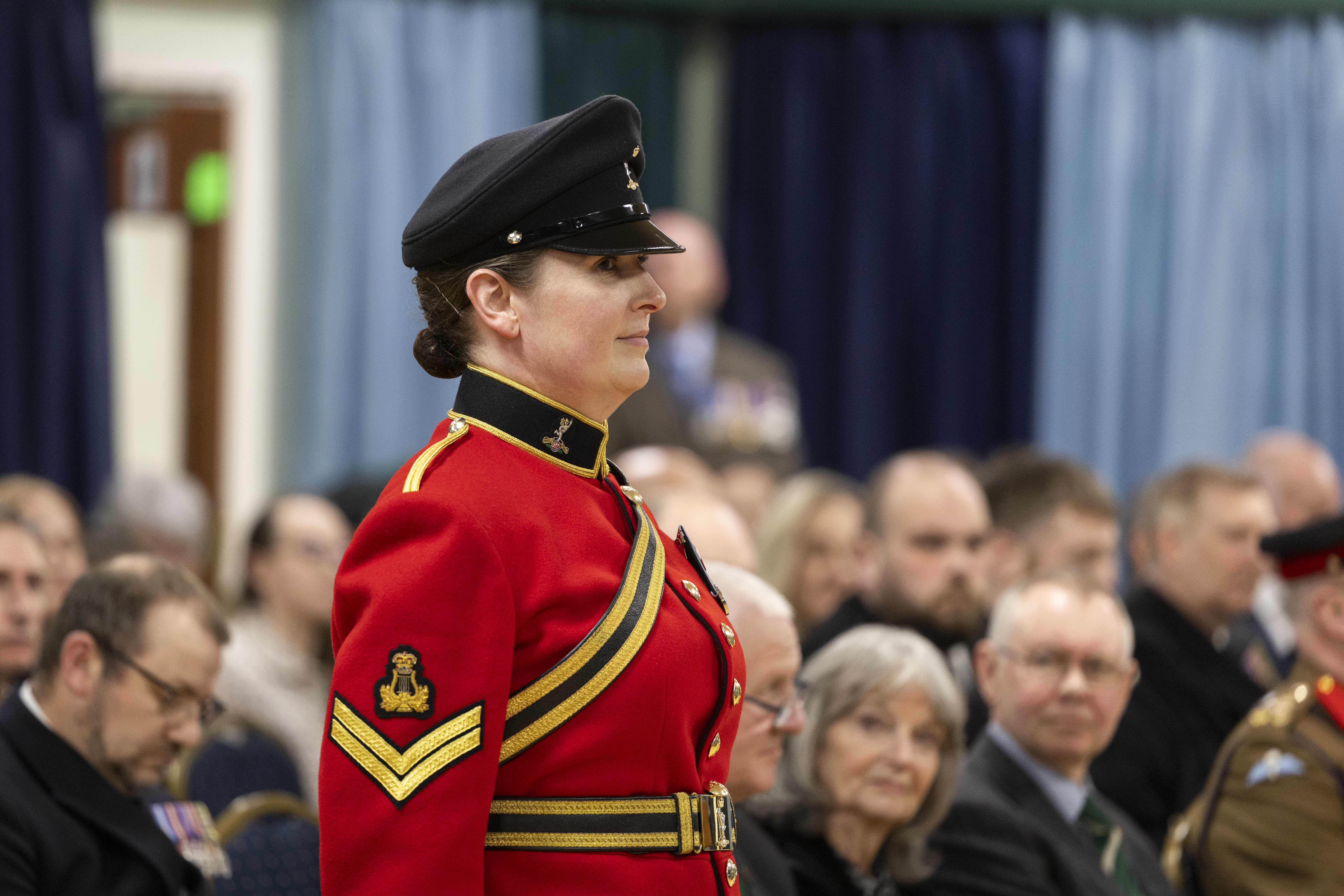 A woman in a red military uniform and black cap stands confidently at an indoor ceremony. Seated audience members in formal attire watch attentively.