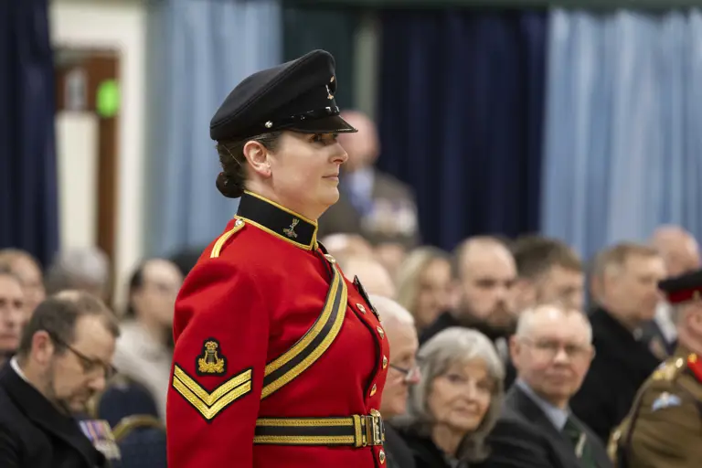 A woman in a red military uniform and black cap stands confidently at an indoor ceremony. Seated audience members in formal attire watch attentively.