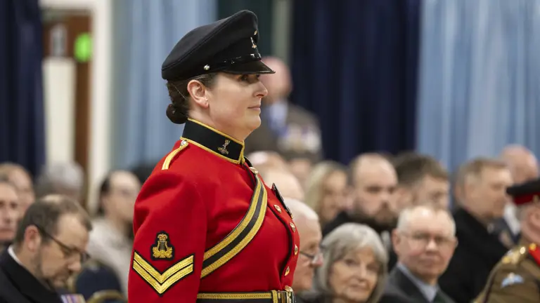 A woman in a red military uniform and black cap stands confidently at an indoor ceremony. Seated audience members in formal attire watch attentively.