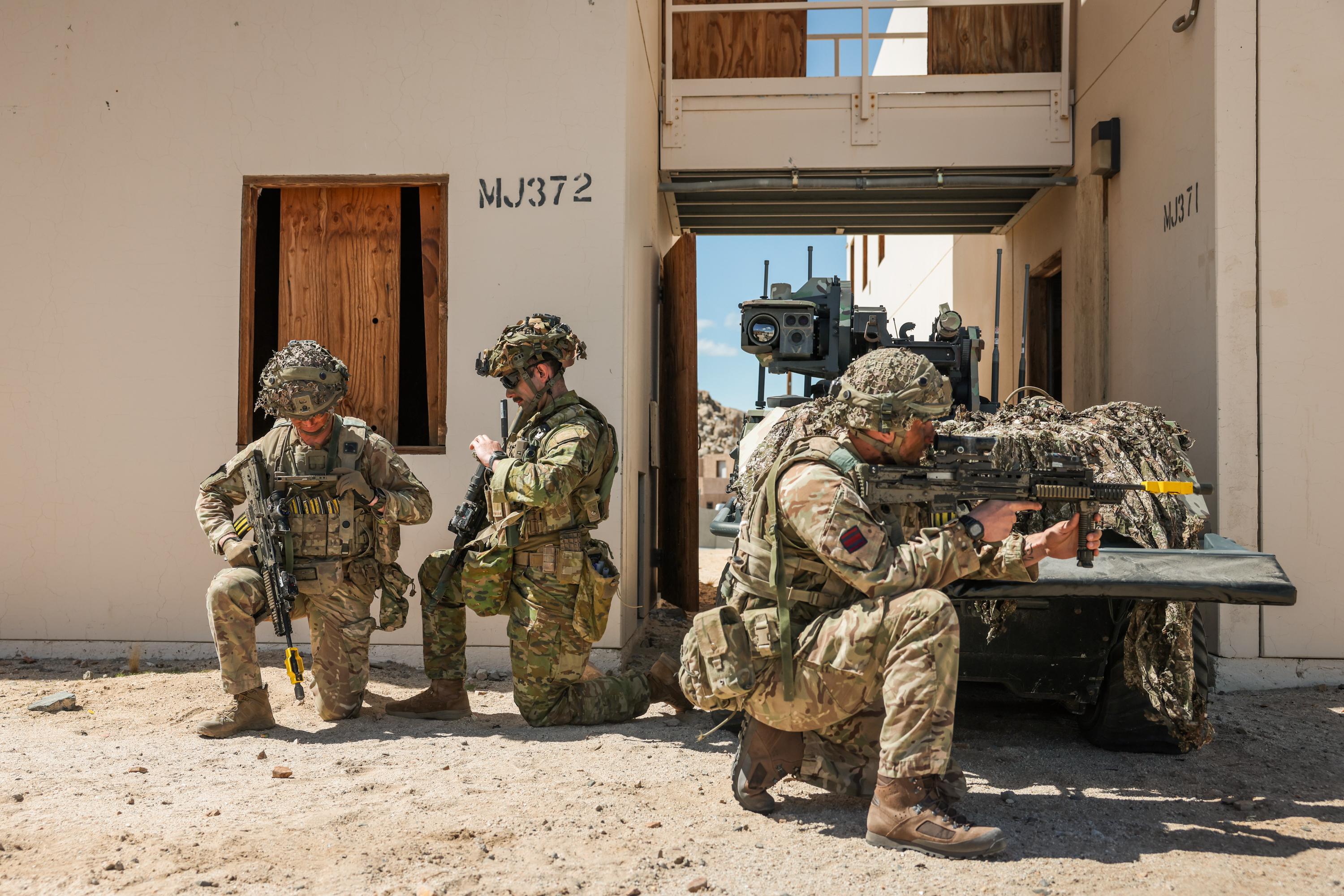 Two British soldiers and an Australian soldier kneel in front of a small green vehicle.