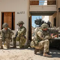 Two British soldiers and an Australian soldier kneel in front of a small green vehicle.