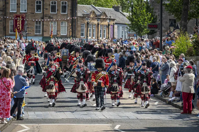 Musicians wearing red tunics and green and blue kilts march along a road.