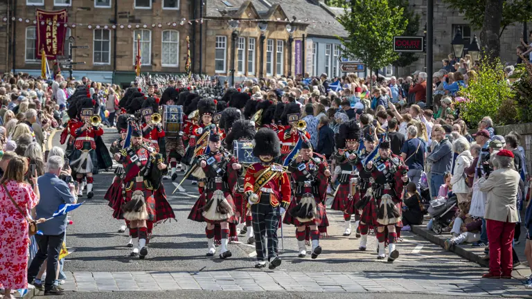 Musicians wearing red tunics and green and blue kilts march along a road.
