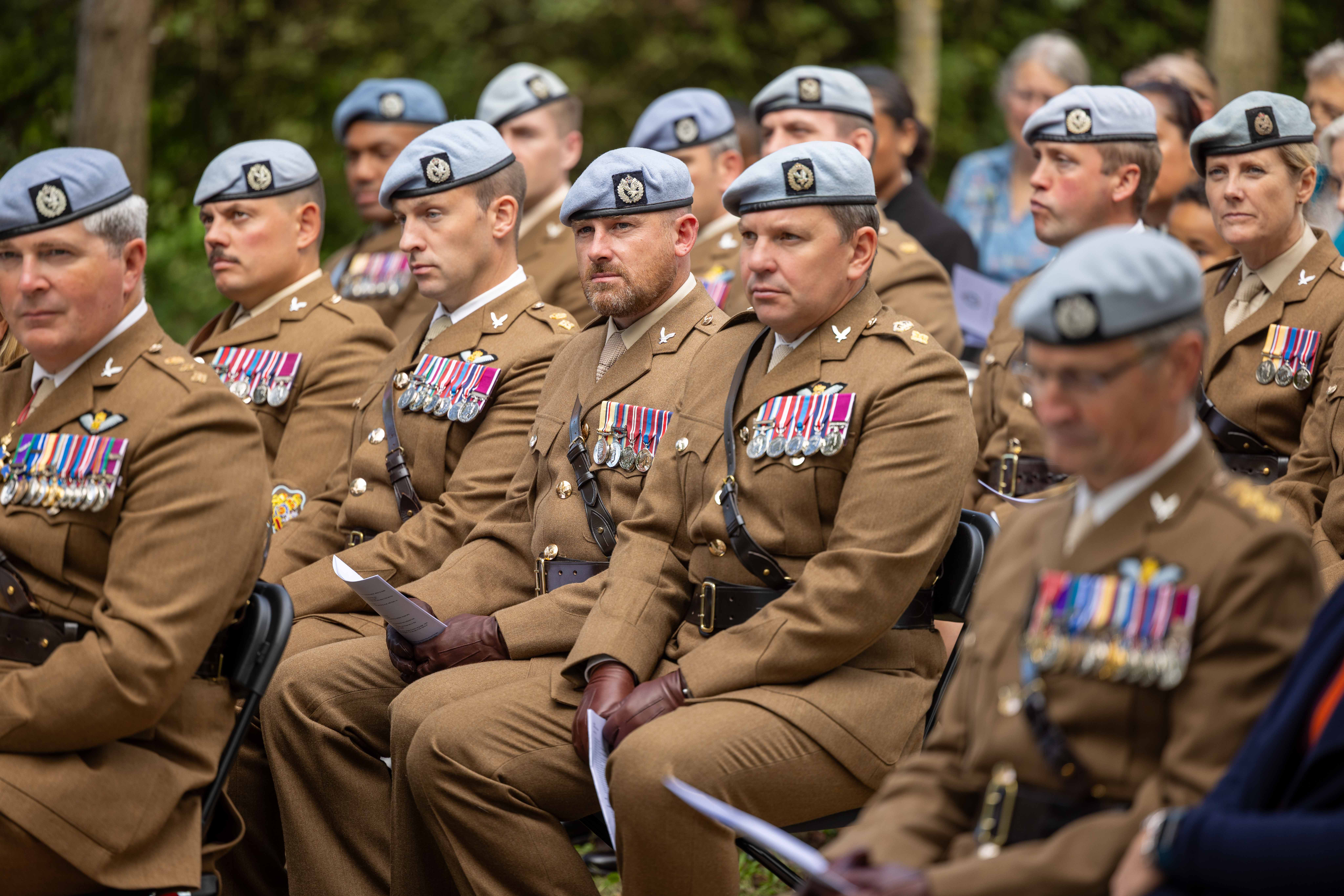 A group of uniformed military personnel is seated outdoors. They are wearing brown uniforms with various medals and light-colored berets. The setting appears formal, with several attendees in the background, surrounded by greenery.
