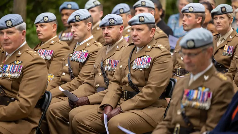 A group of uniformed military personnel is seated outdoors. They are wearing brown uniforms with various medals and light-colored berets. The setting appears formal, with several attendees in the background, surrounded by greenery.