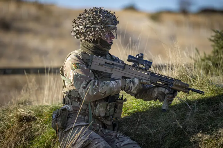 A man in uniform is pictured holding a weapon against in a grassy landscape.