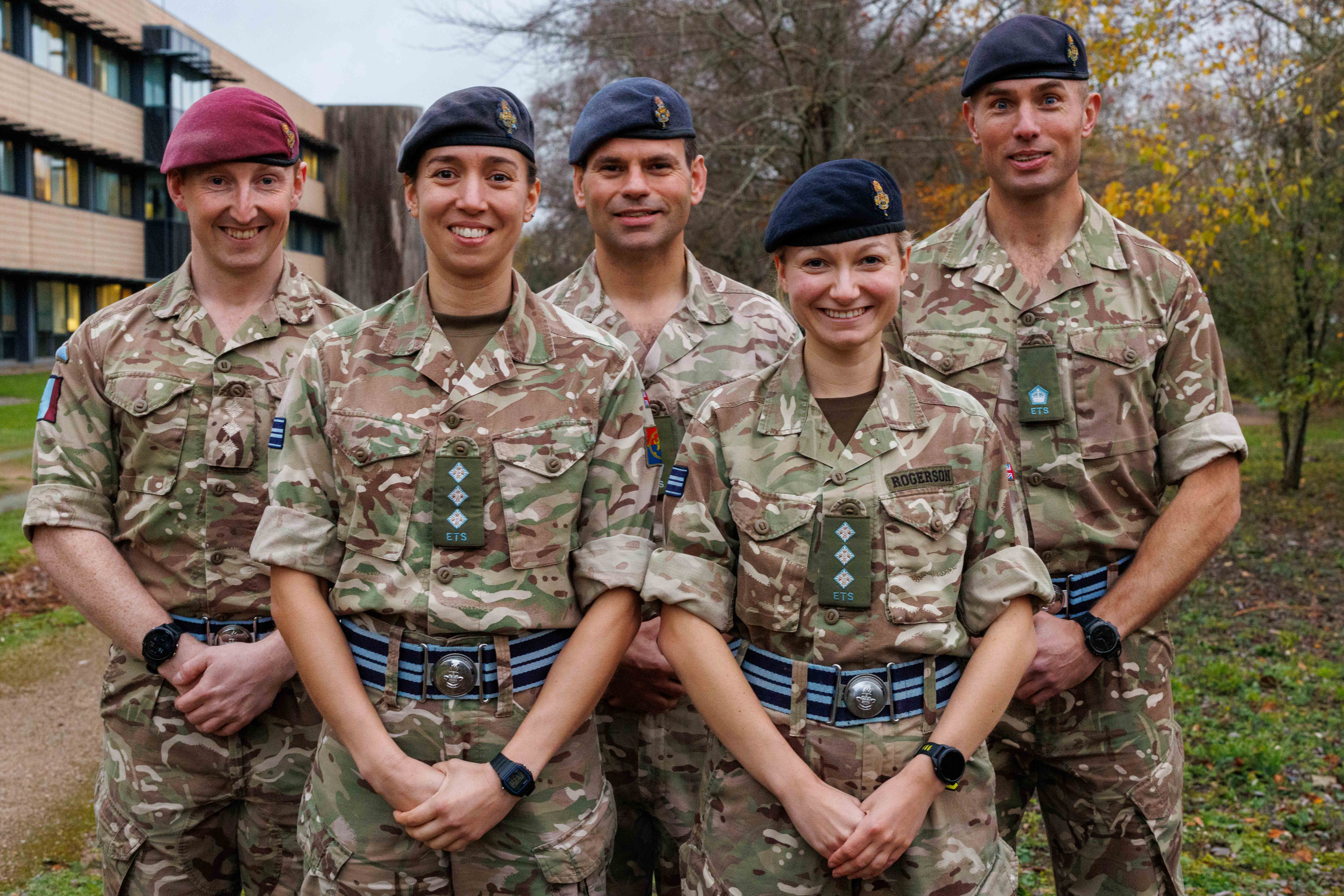 Five soldiers in camouflage uniform pose for a photo outside with a building in the background.