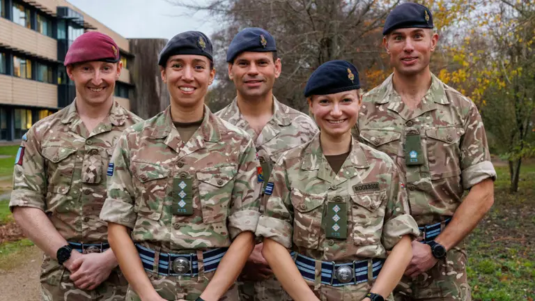 Five soldiers in camouflage uniform pose for a photo outside with a building in the background.