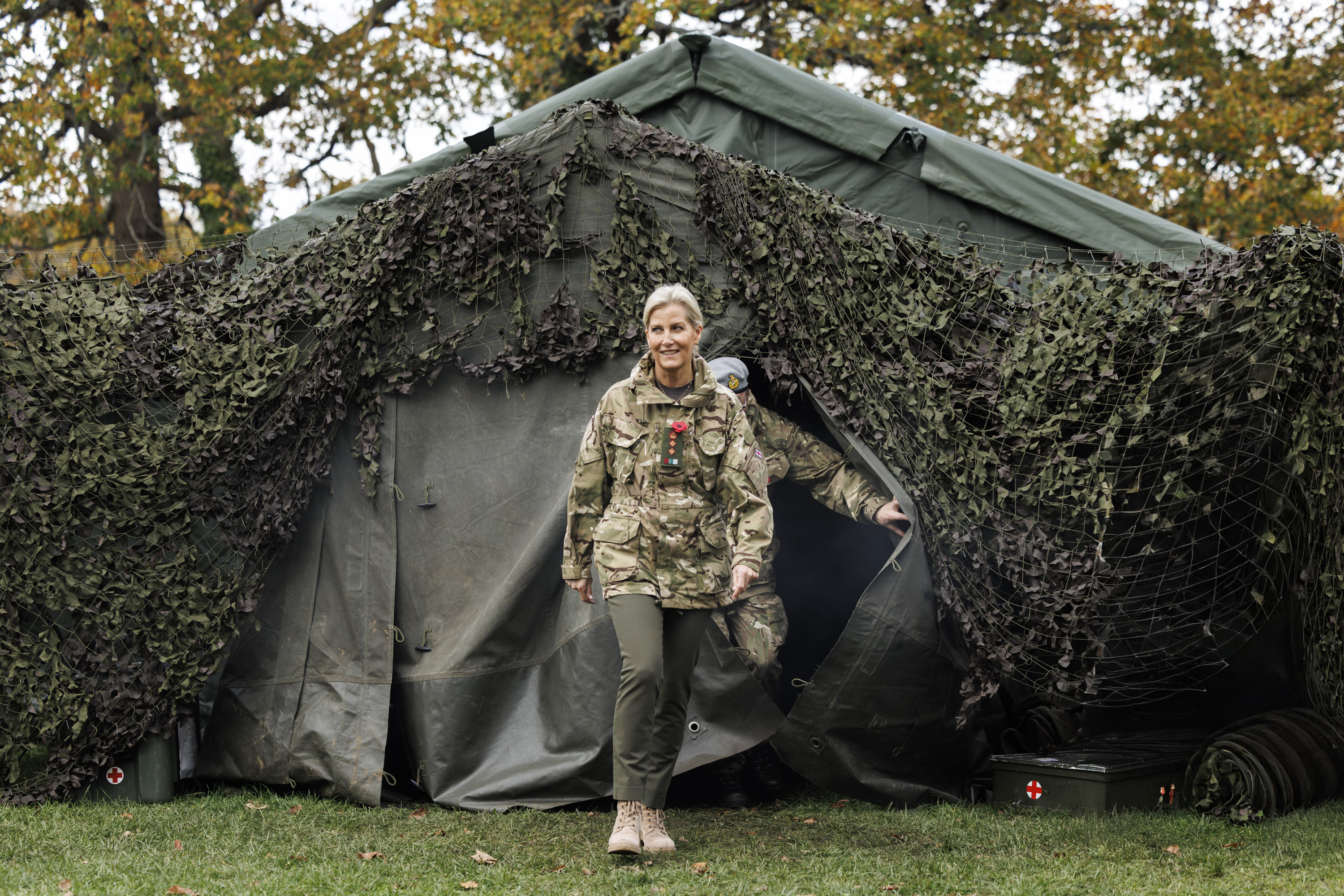 Royal in camouflage uniform walking out of a large military tent covered with netting in a wooded area during autumn.