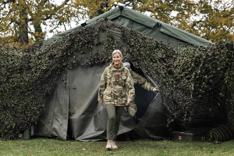 Royal in camouflage uniform walking out of a large military tent covered with netting in a wooded area during autumn.