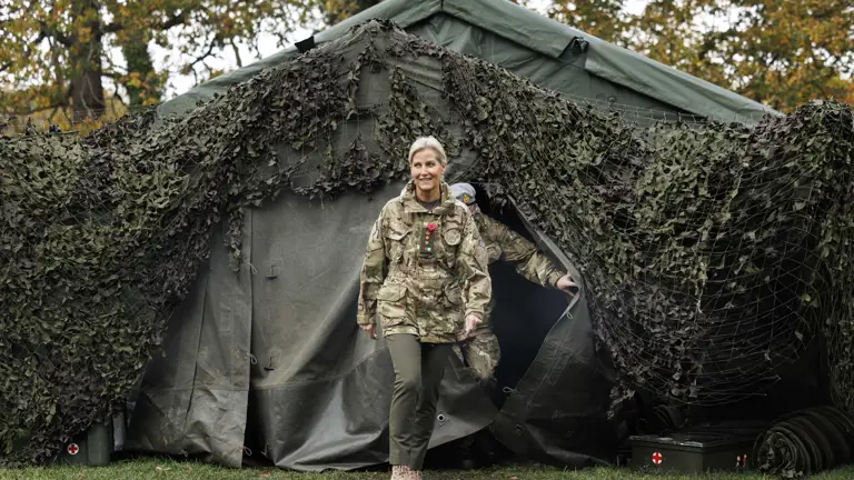 Royal in camouflage uniform walking out of a large military tent covered with netting in a wooded area during autumn.