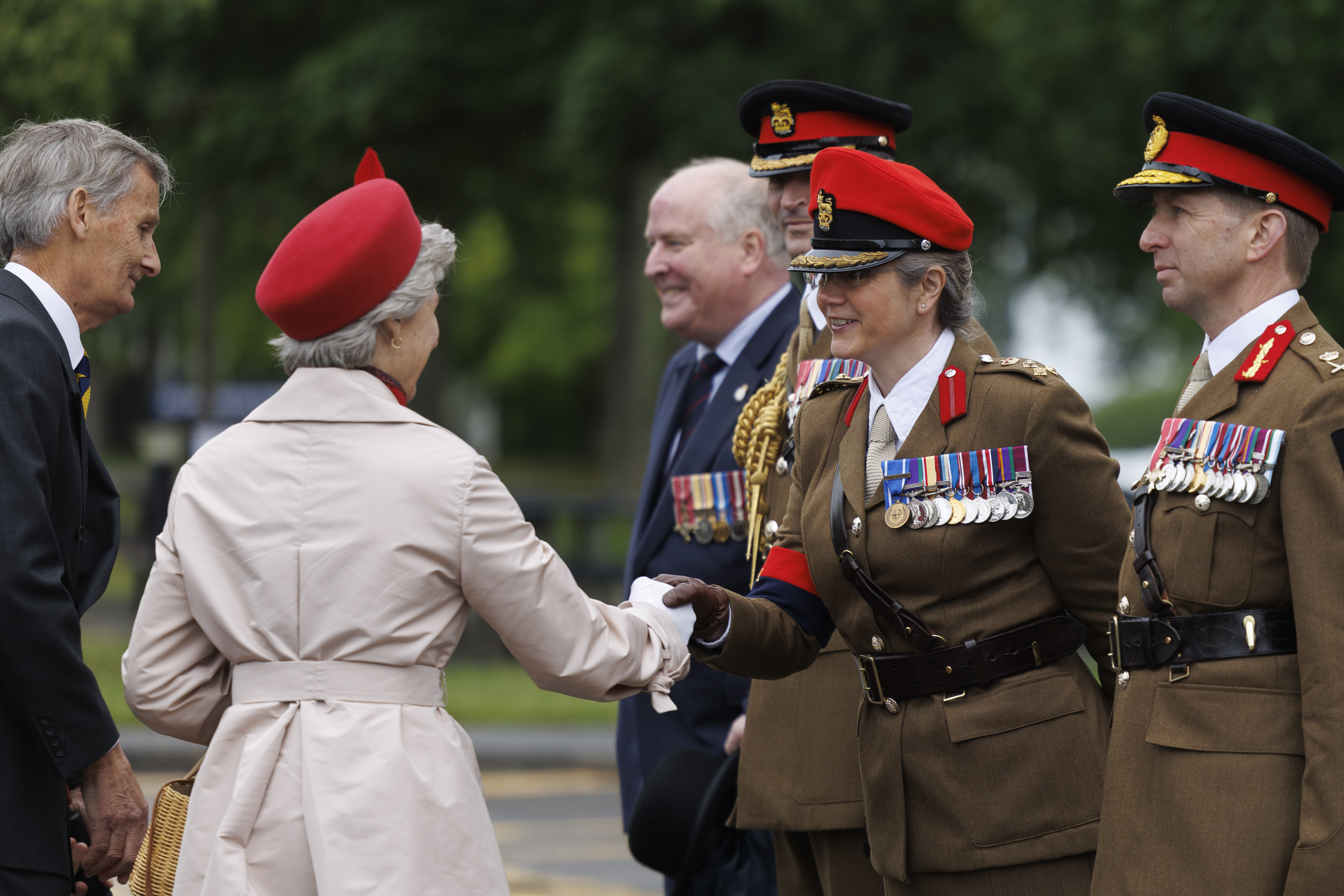Soldiers stood in line next to each other wearing their brown smart uniforms with red caps on their heads. one is shaking the hand of a women wearing a cream coat and a red hat. Two men in blue suits are seen next to them.