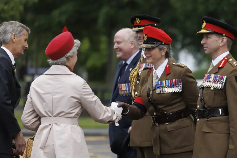 Soldiers stood in line next to each other wearing their brown smart uniforms with red caps on their heads. one is shaking the hand of a women wearing a cream coat and a red hat. Two men in blue suits are seen next to them.