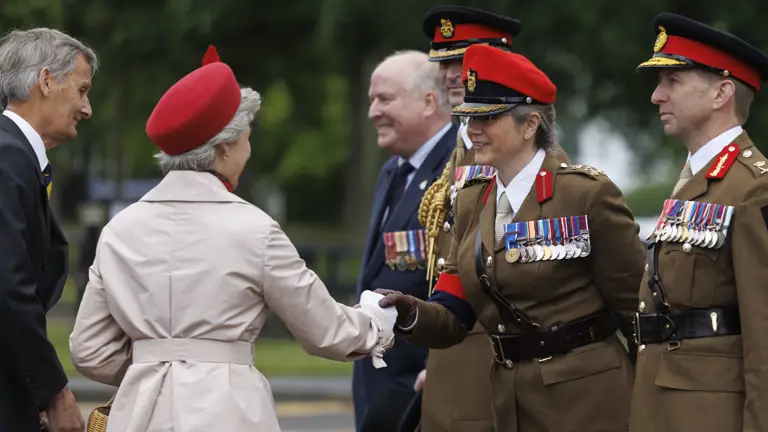 Soldiers stood in line next to each other wearing their brown smart uniforms with red caps on their heads. one is shaking the hand of a women wearing a cream coat and a red hat. Two men in blue suits are seen next to them.