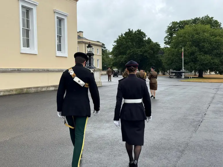 Two individuals in formal military attire walk side by side on a paved area near the Royal Military Academy Sandhurst.