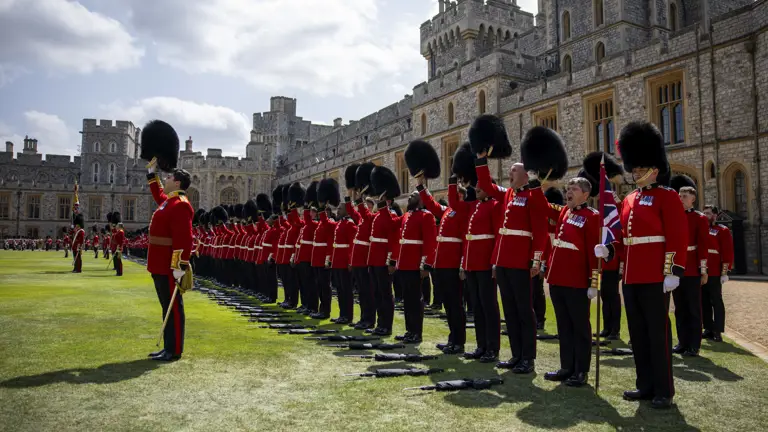 A line of soldiers in red tunics lift their bearskin hats into the air.