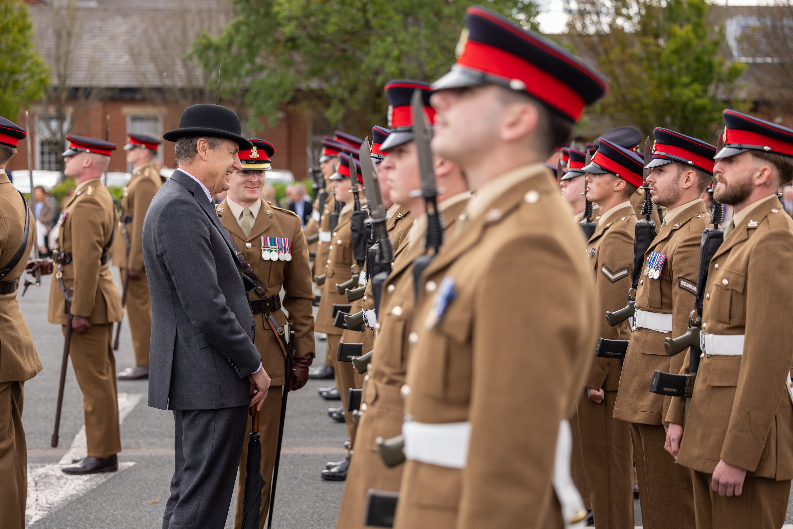 A person in a formal suit and bowler hat interacts with uniformed soldiers standing in formation. The soldiers wear brown uniforms with red and black peaked caps. They are outdoors, with trees and a building visible in the background.