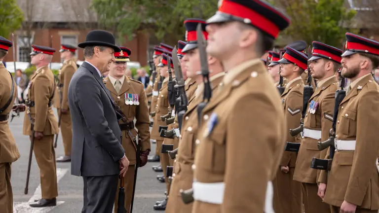 A person in a formal suit and bowler hat interacts with uniformed soldiers standing in formation. The soldiers wear brown uniforms with red and black peaked caps. They are outdoors, with trees and a building visible in the background.